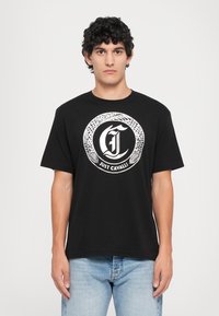 Hombre con camiseta negra con diseño de serpiente circular blanca y texto "Just Cavalli", combinado con jeans de color azul claro, de pie frente a un fondo blanco.