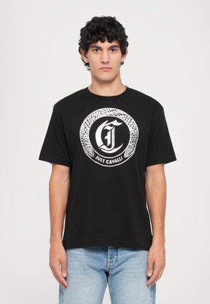 Uomo che indossa una maglietta nera con un design di serpente circolare bianco e il testo "Just Cavalli", abbinata a jeans blu chiaro, in piedi contro uno sfondo bianco.