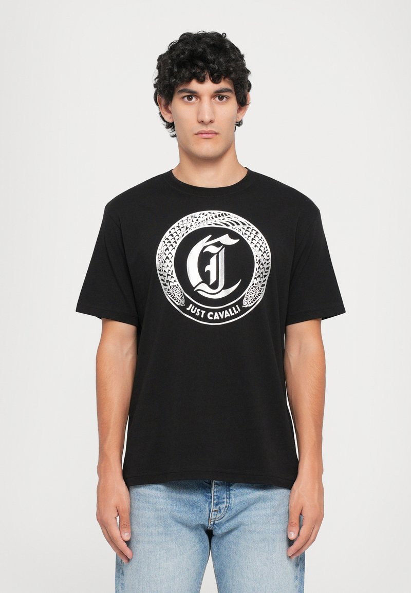Hombre con camiseta negra con diseño de serpiente circular blanca y texto "Just Cavalli", combinado con jeans de color azul claro, de pie frente a un fondo blanco.