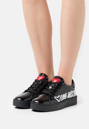 Svarta patentläder sneakers med rund tå, med ett hjärtaccent på tungan, vit text och en svart plattformssula.