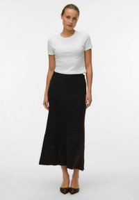Femme se tenant debout, portant un haut blanc à manches courtes crop, une jupe longue noire à côtes et des ballerines brunes à bout pointu, devant un fond blanc uni.