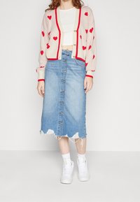 Cardigan lavorato a maglia con motivo a cuore rosso, sfondo crema, chiusura con bottoni, abbinato a una gonna a trapezio in denim sfrangiato, indossato con sneakers bianche.