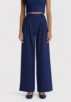 Donna che indossa pantaloni blu navy a vita alta e gamba larga con pieghe, chiusura con bottone e zip, abbinati a scarpe basse blu navy.