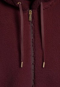 Giacca in pile bordeaux con superficie testurizzata, zip dorata e lacci coordinati. Presenta un colletto alto e una vestibilità casual.