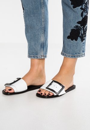 Białe klapki z dużą czarną klamrą, z płaską czarną podeszwą. Noszone z jasnoniebieskimi dżinsami zdobionymi czarną haftowaną florą.