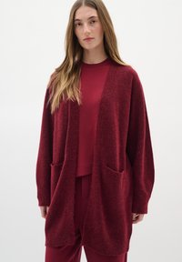 Pikkade heledate pruunide juustega naine kannab laia, tekstuuriga burgundi värvi kardigani, mis on sobitatud burgundi värvi pluusi ja pükstega valge tausta ees.