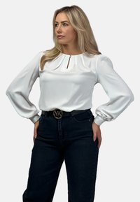 Biała bluzka z plisowanym dekoltem, długimi rękawami w kształcie balonów i detalem w postaci dziurki, połączona z ciemnoniebieskimi dżinsami o wysokim stanie i czarnym paskiem z metalowym logo.