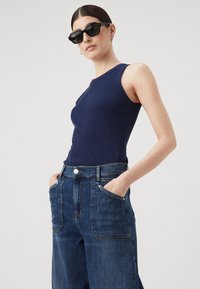 Mornarsko rebrasta majica bez rukava u kombinaciji s visokim tamnoplavim trapericama širokog kroja. Model nosi crne kutne sunčane naočale.