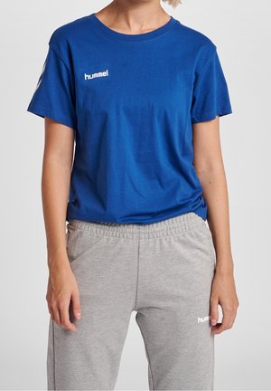 Persoon die een blauw hummel T-shirt en grijze hummel trainingsbroek draagt, staat met ontspannen handen langs het lichaam.