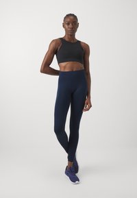 Fekete sportmelltartó mély kivágással, navy magas derekú leggings-szel párosítva. A modellen navy sportcipők vannak. Egységes, sima textúra.