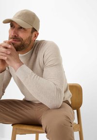Hellbeige Kappe, strukturierter Strickpullover, lockere Passform und braune Hose. Die Person sitzt auf einem Holzstuhl vor einem schlichten weißen Hintergrund.