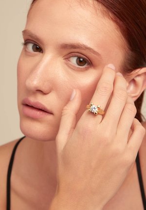 Anillo de oro con una gran gema central clara, rodeada por cuatro acentos esféricos, llevado en una mano cerca de una cara.