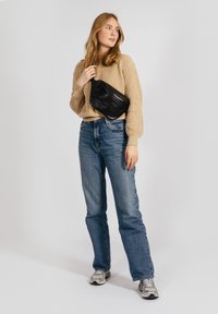 Borsa a tracolla in pelle nera con texture arricciata, indossata sopra un maglione lavorato a maglia beige e jeans a vita alta blu. Sfondo chiaro.