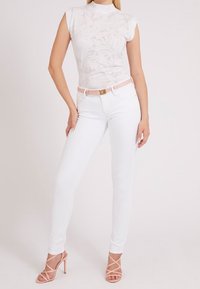 Hvide tætsiddende jeans og en hvid mønstret top med korte ærmer. Sættet har et lyserødt bælte og strop sandaler med nude høje hæle.