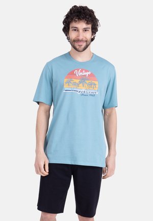 Homme aux cheveux bouclés portant un t-shirt bleu clair avec un motif vintage de coucher de soleil et de palmier, associé à un short noir, debout et souriant.