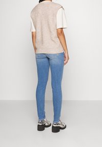 Beige stickad ärmlös topp över en vit T-shirt, matchad med blå skinny jeans och silverfärgade klackskor, vy från ryggen.