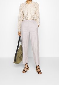 Camicia beige con motivo a occhielli e due tasche; pantaloni di lino grigio chiaro; sandali neri con cinturini; borsa verde oliva con tracolla lunga.