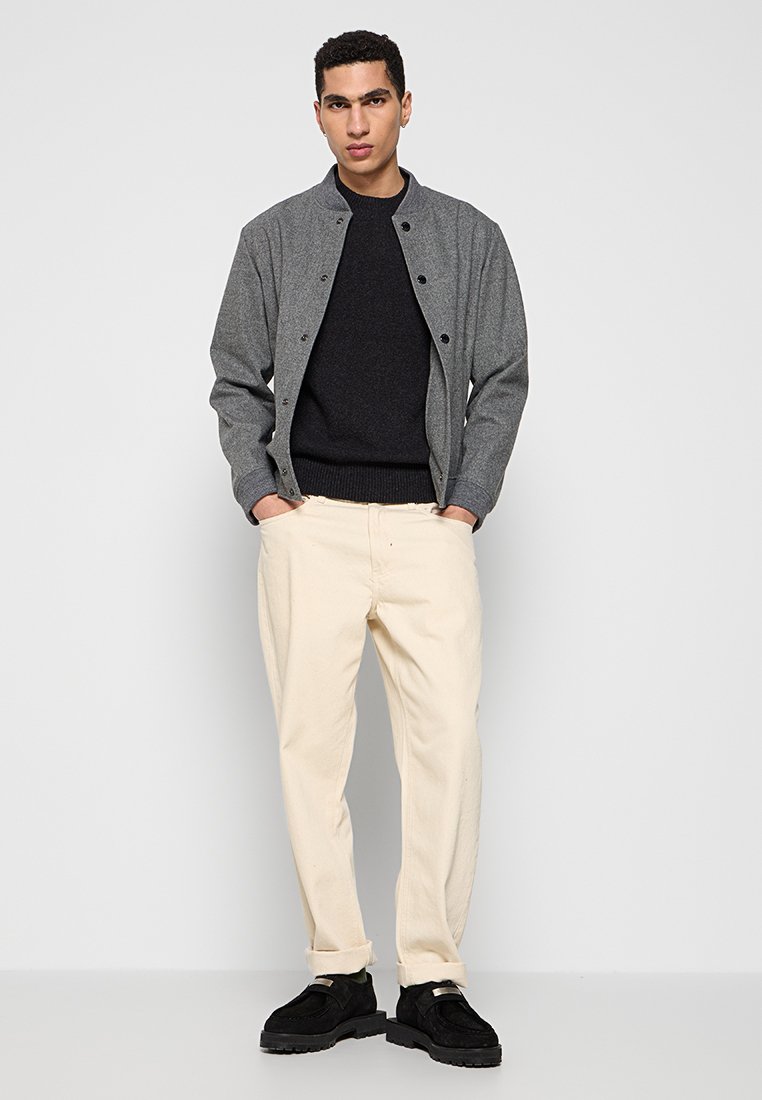 Graue Button-Down-Jacke über einem schwarzen Pullover, beigefarbene Hose und schwarze Schuhe, mit lässigem Schnitt und kontrastierenden Texturen.
