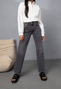 Grå denimjeans kombinerade med en vit långärmad polo. Outfitten har en avslappnad passform, tydlig sömnad och svarta sandaler med remmar.