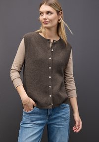 Gilet en maille côtelée marron avec fermeture à boutons, associé à une chemise beige à manches longues. Jean en denim bleu clair. Boucles d'oreilles rondes simples.