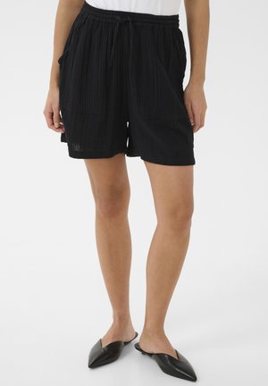 Osoba ubrana w czarne teksturowane szorty z przednimi kieszeniami, biały top i czarne wsuwane buty z ostrym noskiem, stojąca na tle jednolitego, jasnego tła.