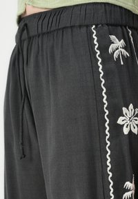 Black fabric pants with an elastic waistband, featuring white embroidered palm and flower designs along the side. Textured and lightweight.