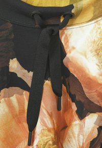 Schwarzer Kordelzug mit Schnürstoppern auf einem bunten, blumenmusterartigen Stoff; bietet warme Töne in Orange, Gelb und Dunkelbraun. Weiche Textur.
