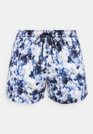 Muž s dlhými dredmi bez trička stojí bosý, oblečený v modro-bielych plavkových šortkách s tie-dye vzorom, ruky v vreckách, na neutrálnej pozadí.