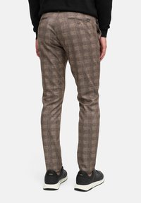 Una persona está de pie de espaldas al espectador, vistiendo pantalones marrones con estampado y zapatillas negras. Se pueden ver dos bolsillos traseros.