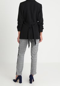 Schwarze Blazer mit einem Bindegürtel in der Taille, kombiniert mit grauen Hahnentritt-Hosen mit schwarzen Seitenstreifen. Trägt dazu navyfarbene Absatzschuhe.