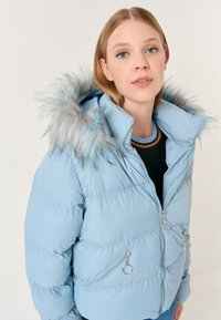 Giacca imbottita blu chiaro con cappuccio in pelliccia, caratterizzata da sfumature di grigio e blu, tasche con zip e dettagli in metallo circolari, e un design trapuntato.