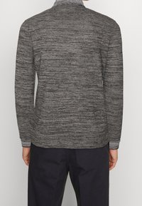 Homme portant un pull en maille grise à manches longues et un pantalon noir, vu de dos sur un fond uni.