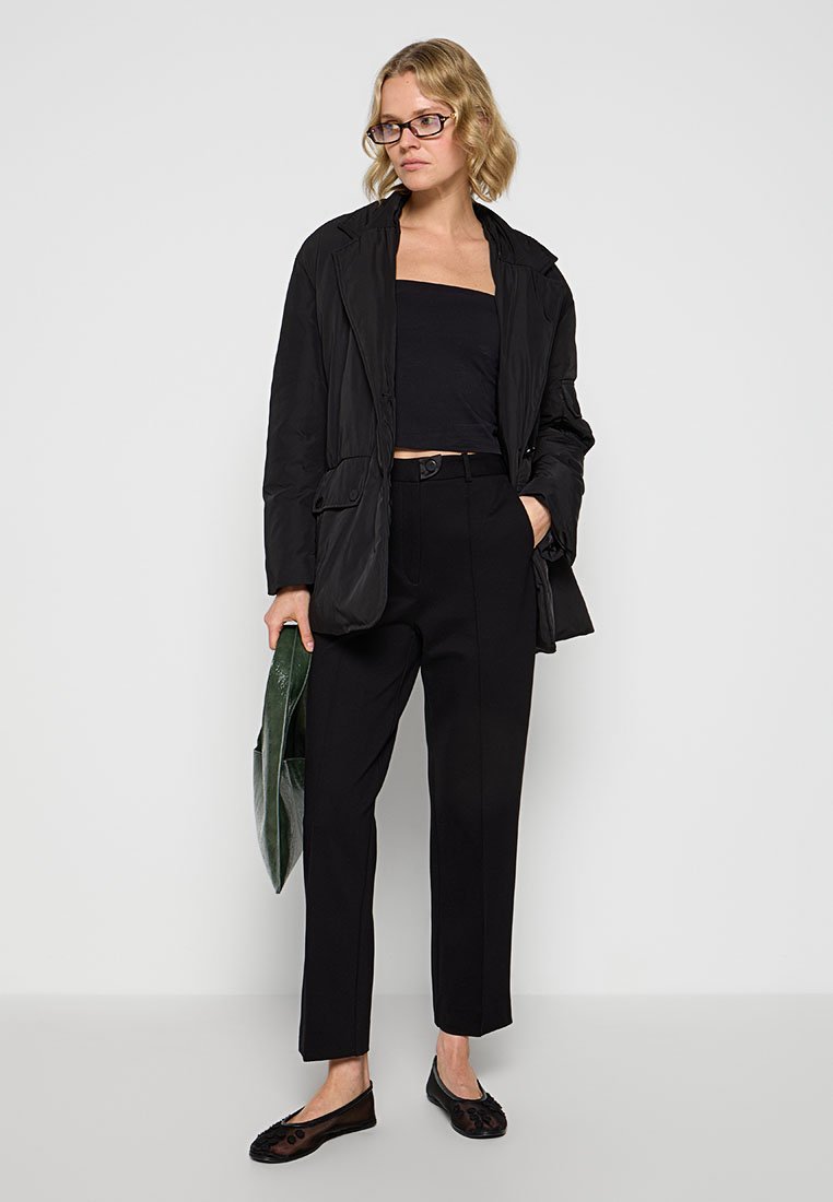 Schwarze gepolsterte Jacke mit tailliertem Gürtel, schwarzes kurzes Oberteil, maßgeschneiderte schwarze Hose und schwarze Ballerinas, während sie eine grüne Tragetasche hält.