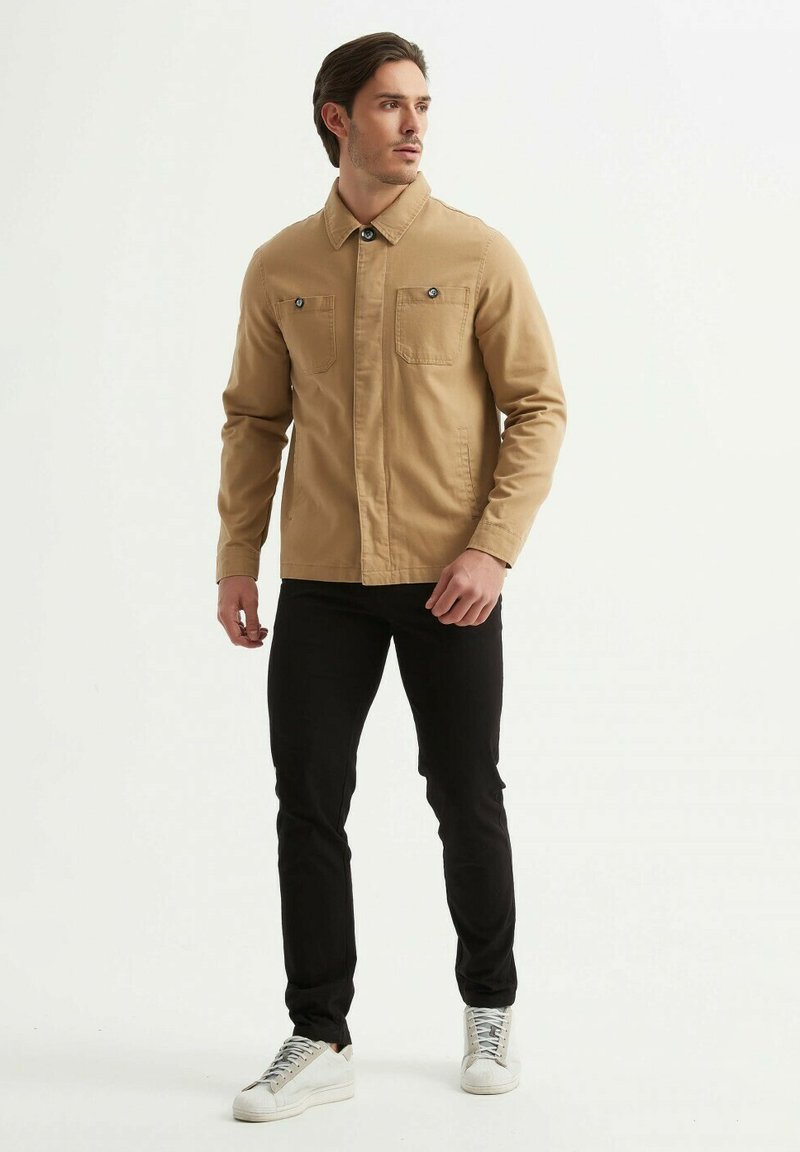 Veste en toile beige avec un col à boutons, deux poches sur la poitrine et un pantalon slim noir. Complété par des baskets blanches et une texture lisse.