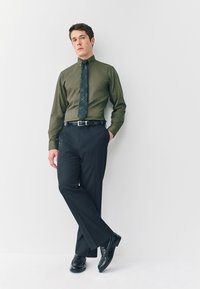 Camisa de botones verde oscuro con corbata de cuadros en tonos azul oscuro y verde; pantalones de vestir negros y zapatos negros brillantes completan el conjunto.