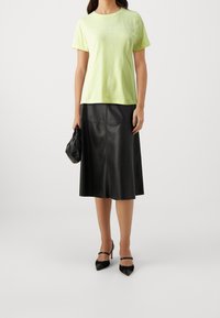 Mujer con una camiseta ligera verde de Tommy Hilfiger, falda midi de cuero negra, tacones de punta negros y sosteniendo un pequeño bolso negro de mano.