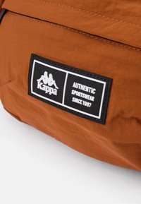 Bruine nylon sportkleding patch met een zwarte rand en witte tekst. Logo bevat twee figuren, met de tekst "Authentic Sportswear Since 1967."