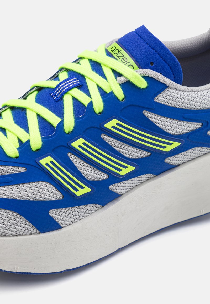 Scarpa da ginnastica blu con parte superiore in rete, lacci verde neon e sovrapposizioni a motivi. Presenta una suola intermedia bianca texturizzata e dettagli blu.