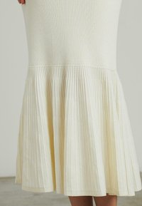 Rippstrickkleid in Off-White mit einem ausgestellten, plissierten Saum. Verfügt über einen taillierten Oberteil und ein strukturiertes Gewebe mit vertikalen Linien.