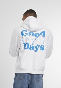 Hvid hættetrøje med blå grafisk tekst "Good Days" og sky-design på bagsiden. Blandingsmateriale af bomuld, afslappet pasform, kengurulomme.