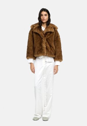 Kobieta o ciemnych włosach, ubrana w brązową kurtkę z futra ekologicznego, białą koszulę na guziki, luźne białe spodnie i jasnoszare buty, stojąca na białym tle.