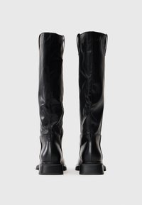 Par de botas de couro pretas altas em pé com saltos baixos, vistas de trás sobre um fundo neutro.