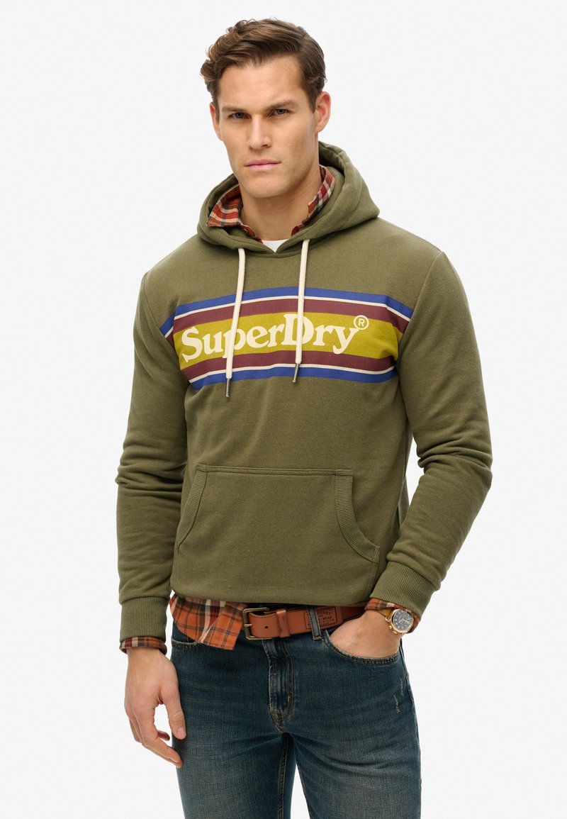 Sudadera de color verde oliva con un bolsillo frontal, con un logo horizontal colorido. Combinada con una camisa de cuadros por debajo y jeans de mezclilla.