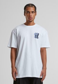 Balta medvilninė t-shirt marškiniai su laisvu pjūviu, priekyje kairėje krūtinės pusėje yra mėlynas logotipas, apvalus iškirpimas ir trumpomis rankovėmis.