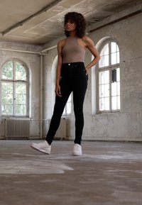 Postava ve béžovém body a černých džínách s vysokým pasem, doplněná o bílé tenisky, stojí v industriálním prostoru s velkými okny.