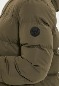 La chaqueta acolchada de color verde oliva presenta una textura suave, un diseño acolchado y un parche redondo negro en la manga izquierda.