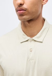 Gros plan sur un homme portant un polo beige clair avec deux boutons fermés, montrant son cou, sa mâchoire et ses lèvres.