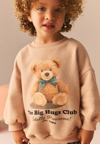 Beżowy sweter z grafiką pluszowego misia w zielonej kokardzie. Tekst brzmi "Klub Wielkich Przytulaków" oraz "Całkowicie Zajefajnie!" na czarnym tle.