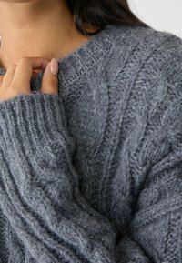 Pull gris en maille torsadée avec poignets côtelés et motifs texturés, mettant en valeur un tissu doux. Gros plan sur le détail de l'encolure et de la manche.