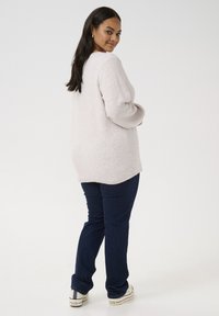 Pulover tricotat de culoare roz deschis, cu o strălucire subtilă, croială lejeră și margine ondulată, asortat cu blugi de culoare albastră închis și teniși albi, văzut din spate.